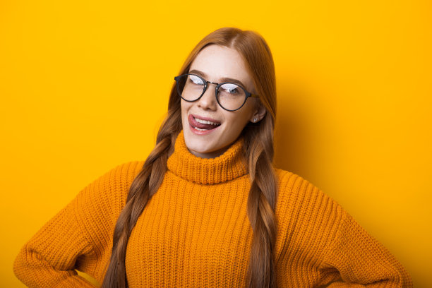 白种女孩红头发和雀斑透过她的眼镜，在黄色背景下摆姿势，穿着毛衣图片下载
