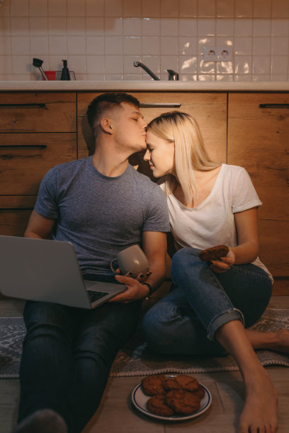 竖版照片：一对白人夫妇在地板上，在厨房里一起用电脑喝热茶图片下载