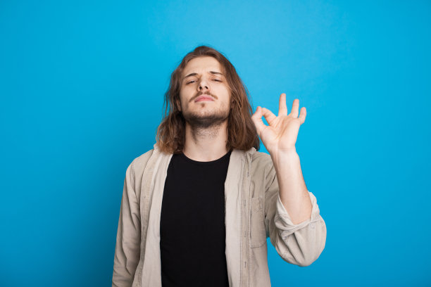 一名蓄着胡须的长头发白人男子在蓝色背景下自信地摆姿势，打着“ok”的手势图片下载