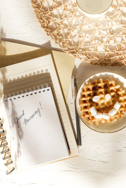 棉花糖华夫饼微笑和笔记本写着早上好在白色的纹理桌上。平面构成。图片下载