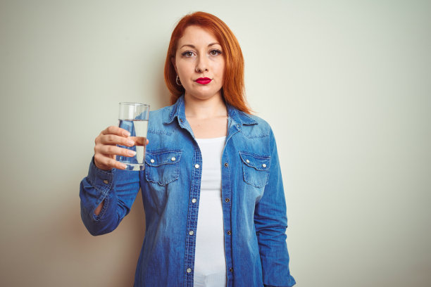 年轻美丽的红发女人喝一杯水在白色孤立的背景与自信的表情在聪明的脸上思考认真图片下载