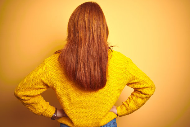 美丽的红发女人穿着冬季毛衣站在孤立的黄色背景站向后看远离与手臂在身体上图片下载