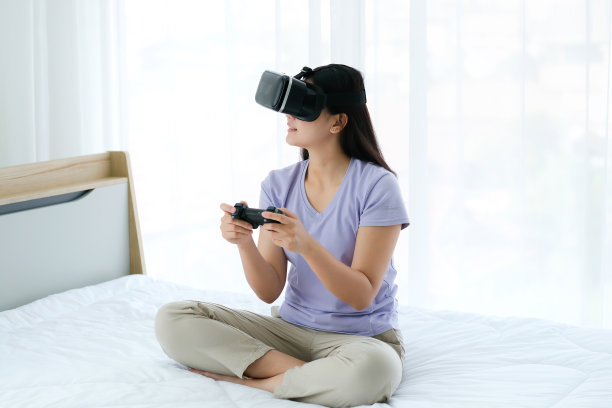 亚洲女性戴着虚拟现实眼镜，坐在卧室的床上。手持操纵杆，使用VR眼镜玩游戏图片下载