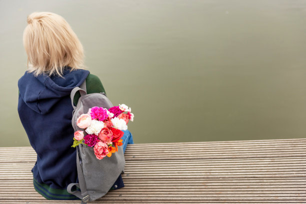 年轻金发梦幻女子在河边拿着一束鲜花，灰色背包的背影照片，浪漫氛围图片下载