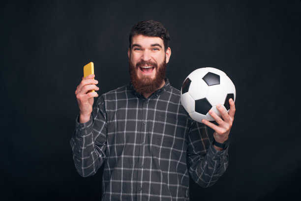 一个长着胡子的快乐的年轻人正拿着手机和一个足球靠近黑色的墙壁。图片下载