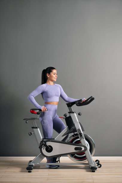 一个漂亮的运动年轻女子身穿运动服，骑着自行车在体育馆的灰色墙壁上。图片下载