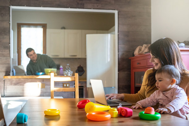 居家厨房里的家庭：妈妈在桌旁用电脑工作，女儿在她腿上玩玩具，爸爸做午饭——千禧一代与孩子的亲密时刻图片下载