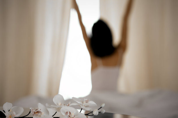 醒来后在床上伸展双手的女人，阳光照射。一个黑发女人在睡了个好觉后，快乐而放松地开始新的一天，背影。新的一天和快乐的周末的概念图片下载