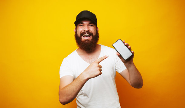 快乐的大胡子嬉皮士指着黄色背景上的智能手机图片下载