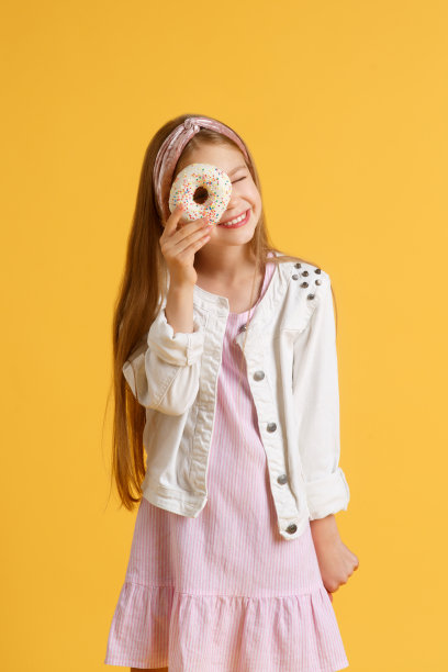 疯狂快乐的金发女孩戴着生日帽微笑，玩得很开心，看着她眼睛上的两个红色甜甜圈。糖果。黄色工作室背景图片下载