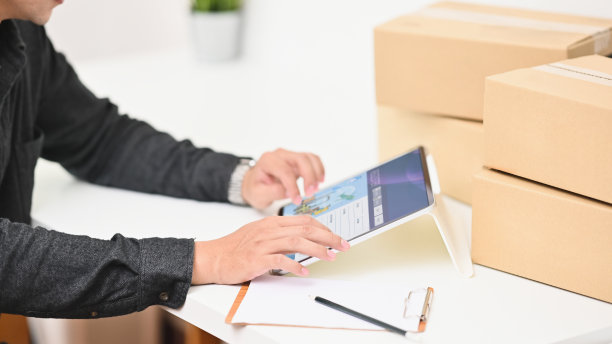 这是一幅年轻企业主坐在现代办公桌旁使用平板电脑的照片，周围是一堆纸箱，背景是舒适的客厅。图片下载