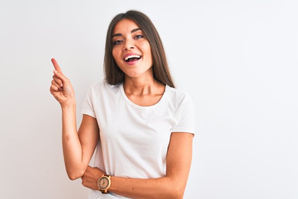 年轻美丽的女人穿着休闲t恤站在孤立的白色背景上，脸上带着大大的微笑，用手指指着一边看着相机。图片下载