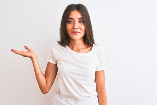 年轻美丽的女人穿着休闲t恤站在孤立的白色背景微笑着提出和指向与手掌看着相机。图片下载