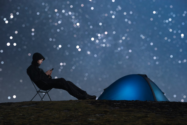 男性游客坐在椅子上使用智能手机。背景是一个发光的帐篷和繁星点点的天空。图片下载