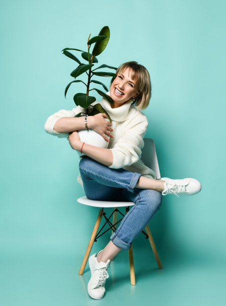 年轻快乐的笑金发女人穿着毛衣，牛仔裤和运动鞋坐在椅子上抱着盆栽榕树图片下载