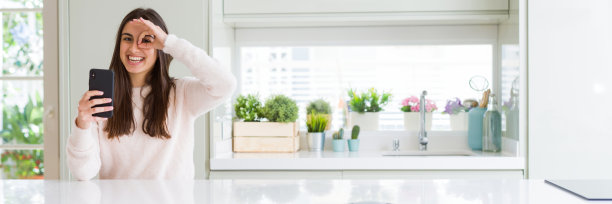 广角图片美丽的年轻女子用智能手机发送信息，快乐的脸，微笑做ok手势，手在眼睛透过手指看图片下载