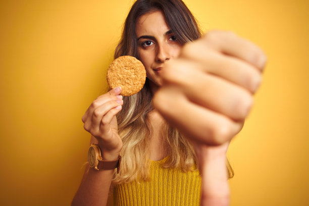 年轻漂亮的女人在灰色背景下吃饼干，面带怒容图片下载