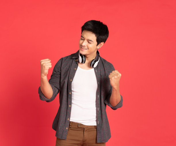 快乐的年轻亚洲学生男子穿着休闲衬衫唱歌和听音乐的手机和无线耳机隔离在红墙和复制空间。图片下载