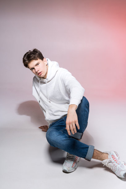 时尚帅气的年轻欧洲男模穿着白色运动衫，蓝色牛仔裤和白色运动鞋在工作室里摆姿势，在粉红色的背景下拍摄商业照片，看着相机图片下载