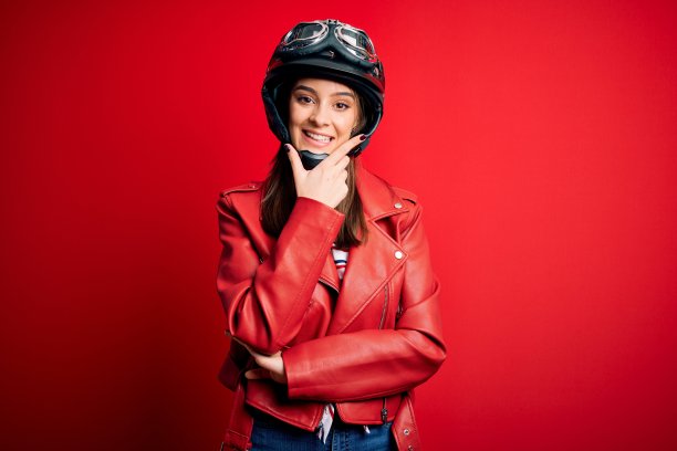 年轻漂亮的黑发女摩托车手戴着摩托车头盔，穿着红色夹克，在镜头前看起来很自信，双臂交叉，手放在下巴上。积极的思考。图片下载