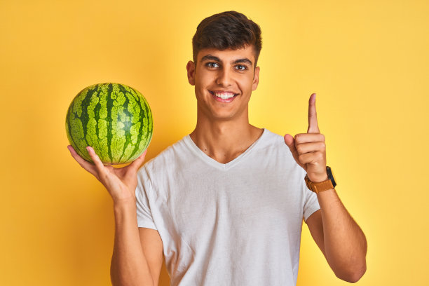 拿着西瓜的年轻印度店主，在黄色背景前惊讶地指着头，想到了主意图片下载