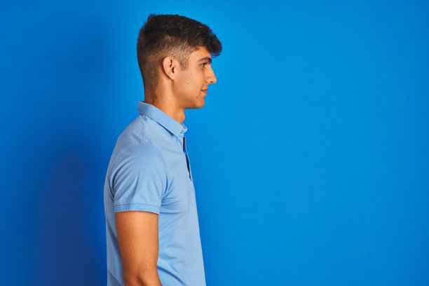 在蓝色背景前，身穿休闲POLO衫的年轻印度男子看向侧面，放松的姿势，面带自信的微笑图片下载