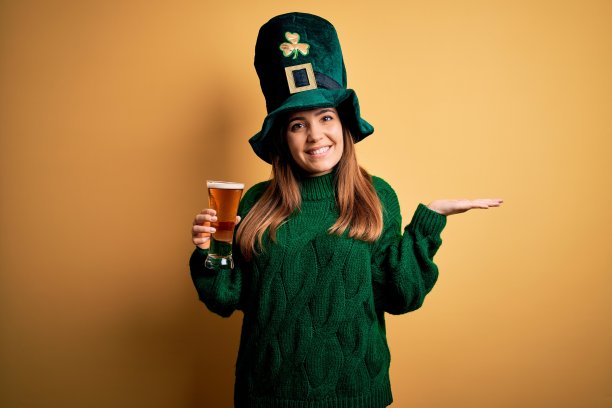 年轻美丽的女人戴着绿帽子喝着一杯啤酒在圣帕特里克节愉快地呈现和指向与手掌看着相机。图片下载