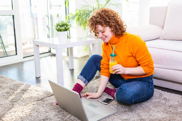 一个红头发的年轻女子在家里喝着橙汁上网。自由职业女性在家用笔记本电脑工作图片下载