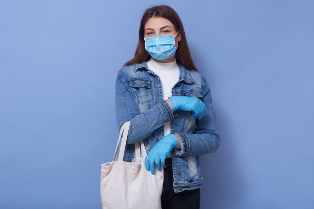 这张照片拍摄的是一个严肃的蓝眼睛的小女孩，她穿着牛仔夹克，戴着防护口罩和手套购物，拿着环保袋，直视镜头，为了安全独自花时间。图片下载