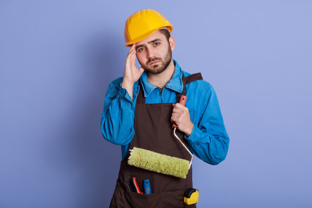 水平的室内图片，有思想的严肃的年轻工人抱着滚轮，工作累了，疲惫的表情，孤立地站在工作室的蓝色背景。工作的概念。图片下载