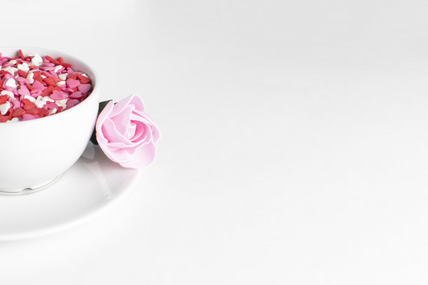 白色的咖啡杯，白色的背景上撒满了心形的五彩纸屑。粉红色的玫瑰是在碟子上选择性的焦点。情人节的概念。图片下载