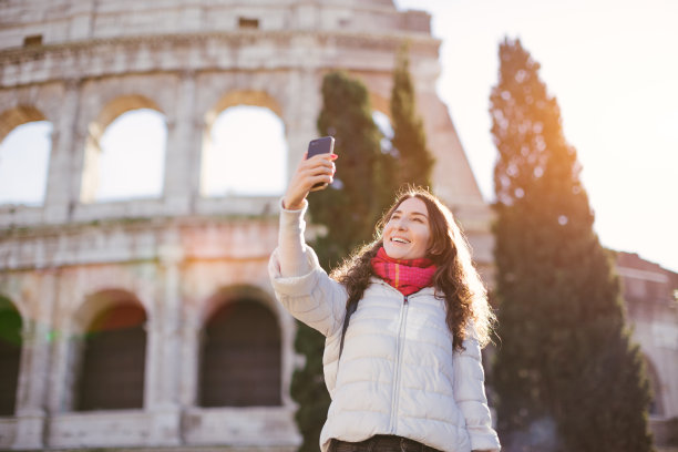年轻美丽的微笑女子与罗马斗牛场(斗牛场)摆pose，并与智能手机自拍-冬季假期和假期在欧洲的概念图片下载