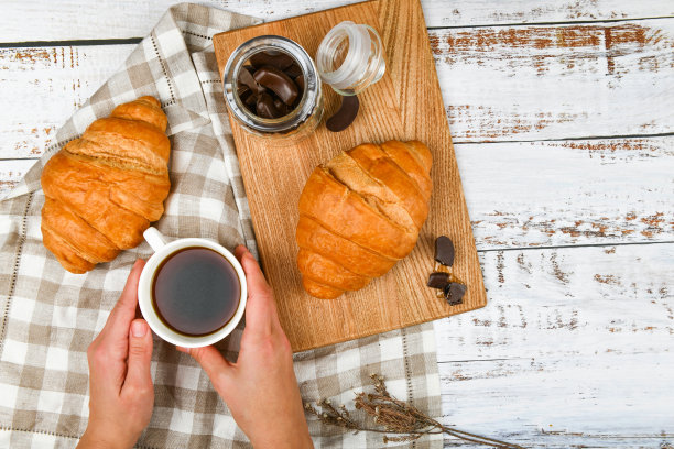 女孩拿着一个咖啡杯。以羊角面包为背景。咖啡和羊角面包。清晨的开始。咖啡杯。俯视图女人的手拿着一杯咖啡和新鲜的法国羊角面包图片下载