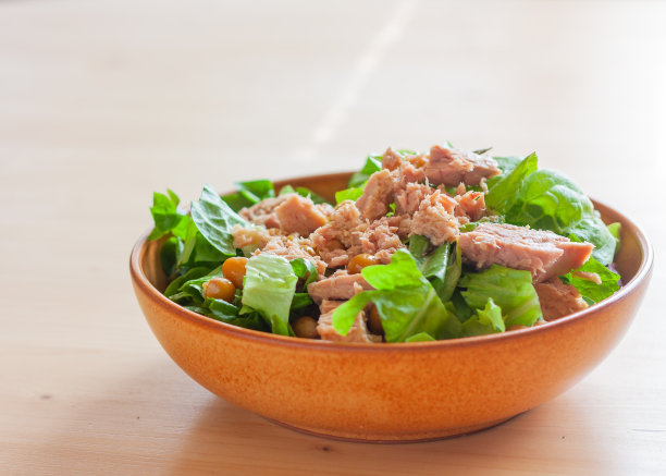 绿色的莴苣、金枪鱼和鹰嘴豆沙拉放在天然的陶瓷盘子里，在木制的光背景上。健康生活和饮食不可或缺的成分图片下载