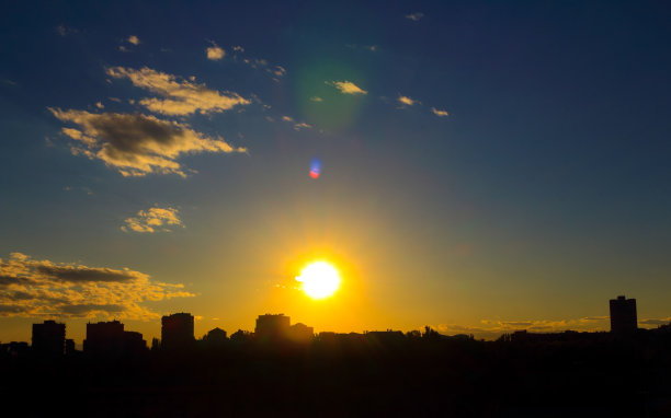 可爱的夕阳，与金色的太阳，与灰白色的云，与蔚蓝的天空，与镜头闪光的背景城市剪影。图片下载