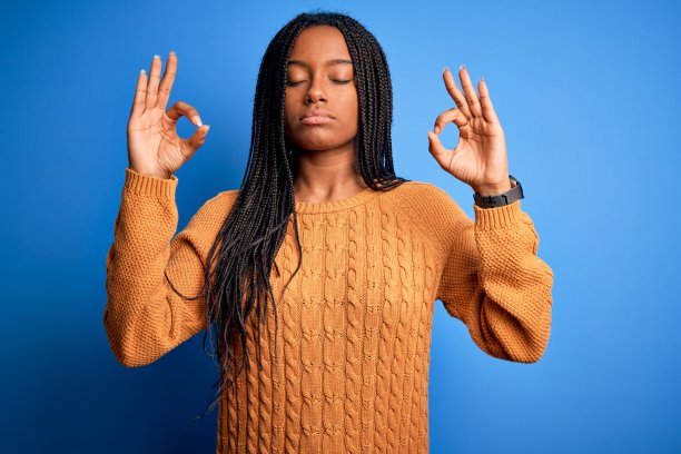 年轻的非洲裔美国妇女穿着休闲黄色毛衣站在蓝色孤立的背景放松和微笑，闭上眼睛做冥想手势与手指。瑜伽的概念。图片下载