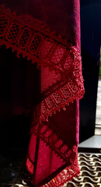 在明亮的阳光下华丽的深红色天鹅绒花边桌布。透明花边边缘的窗帘特写。图片下载