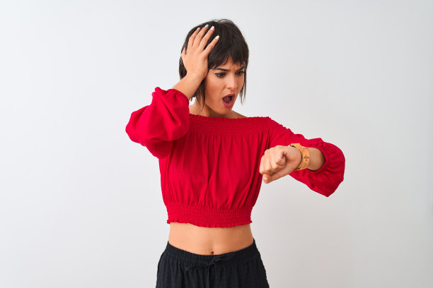 年轻美丽的女子穿着红色的夏季t恤孤立地站在白色的背景下看着手表着急的时间，害怕迟到图片下载