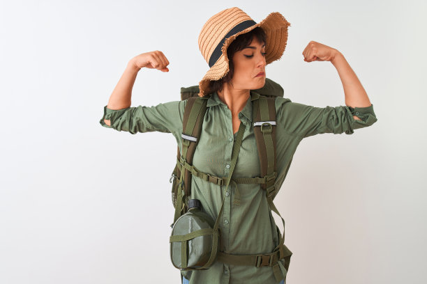 徒步妇女戴着背包帽子和水水壶在孤立的白色背景显示手臂肌肉自豪地微笑。健康的概念。图片下载