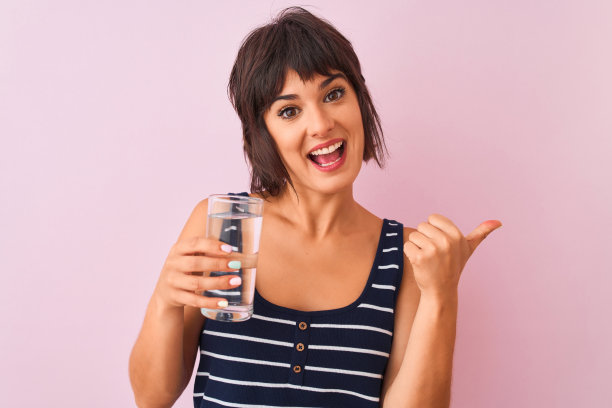 年轻美丽的女人拿着水站在孤立的粉红色背景指向和显示拇指向上到一边与幸福的脸微笑图片下载