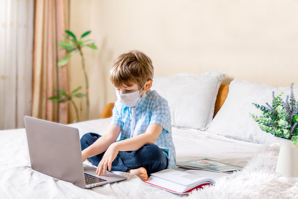 远程学习在线教育。生病的男孩戴着医用口罩在家学习用数码平板电脑笔记本电脑和做学校作业。坐在床上，拿着训练书。图片下载