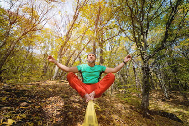 在秋天的森林里，一个长着胡须、赤脚的成年人坐在一条松弛的绳索上冥想。瑜伽和平衡练习。精神健康图片下载