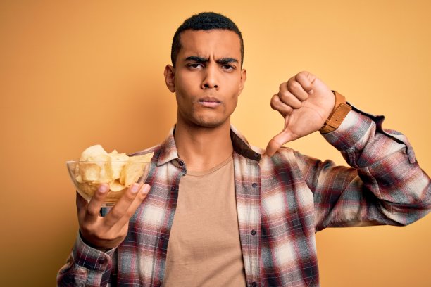 年轻英俊的非裔美国人拿着一碗薯条土豆在黄色的背景与愤怒的脸，消极的迹象显示不喜欢拇指下来，拒绝概念图片下载