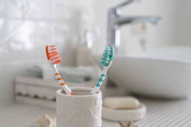 两把彩色的牙刷放在陶瓷玻璃上，背景是洗脸盆和浴室里的其他配件图片下载