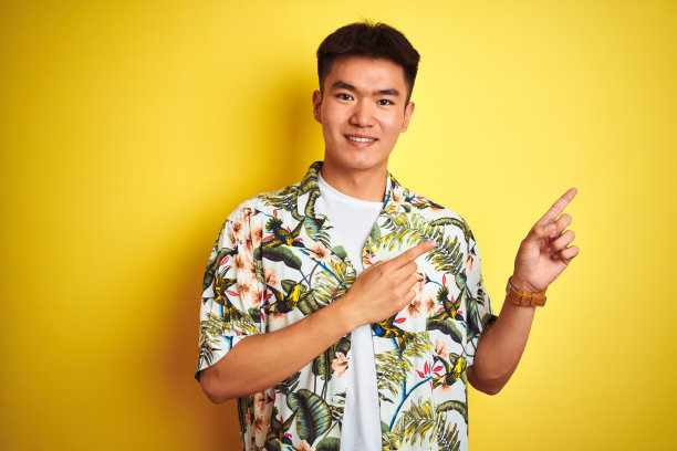 亚洲华人男子度假穿着夏季衬衫在孤立的黄色背景微笑，看着相机指向与双手和手指的一边。图片下载