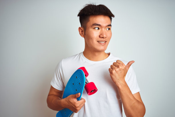年轻的亚洲中国学生男子持有滑板站在孤立的白色背景指向和显示拇指向上到一边与快乐的脸微笑图片下载
