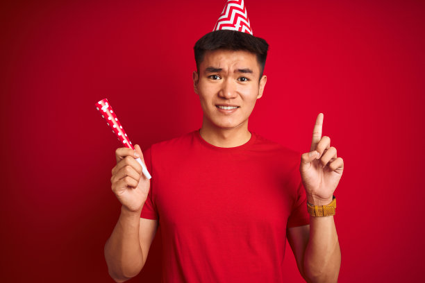 亚洲华人男子在生日庆典上戴着滑稽的帽子在孤立的红色背景下惊讶的一个想法或问题指向幸福的脸，第一图片下载