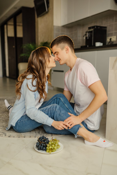 快乐的年轻男女坐在厨房地板上，低头互相沐浴。一对幸福的夫妇在时尚的厨房里。室内照片浪漫的女孩看着男朋友。图片下载