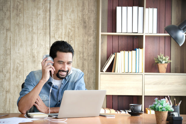 留着胡须的亚洲企业主，耳朵上戴着耳机，一边看着笔记本电脑，一边开心地开着视频会议。温暖的父亲与在国外的女儿视频通话。图片下载
