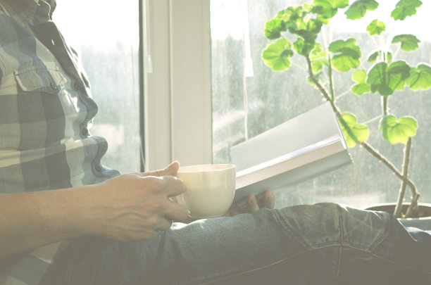 一个穿着格子衬衫和牛仔裤的男人坐在窗台上，拿着一杯咖啡和一个记事本图片下载